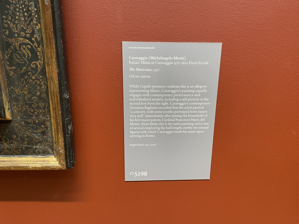 The museum label for Caravaggio’s The Musicians on a red wall beside a gilded picture frame.